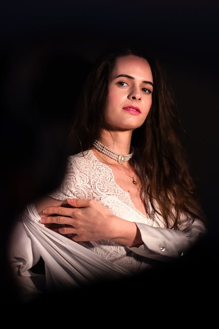 A woman with long, wavy brown hair and light skin is holding a white satin fabric or garment across her chest. She is dressed in elegant attire including a white lace top and a layered pearl necklace with a small pendant. The background is dark, emphasizing her facial features and clothing, which appears to be part of a dress or formal outfit. The lighting highlights her face and upper body, creating a soft, poised appearance suitable for fashion or portrait photography.
