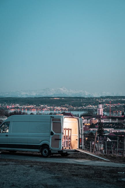 A light grey moving van parked outdoors on a slightly sloped surface, with its side door open revealing stacked packed cardboard boxes and protective foam padding inside. The van is positioned near a 