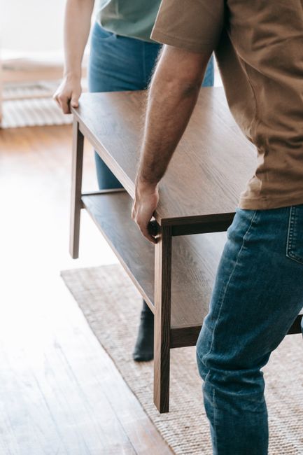A person wearing a brown shirt and blue jeans is lifting a small wooden side table, which has a dark frame and light wooden surface, inside a home. The individual grips the table’s edge, positioning it for transportation as part of a home relocation process. In the background, another person in a brown top and blue trousers stands nearby, observing or assisting. The room features wooden flooring with a woven rug partially visible, and sunlight streams through a window, illuminating the scene. The setting suggests preparation for furniture transport, with attention to careful handling during packing and moving activities. Colliers Wood Movers, a professional removals company, may be involved in such furniture transport and packing services, supporting efficient house removals in the area.